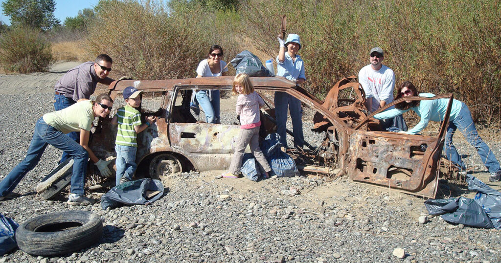 A group of people around a rusted out old station wagon half buried in the gravel creek bed of Cache Creek