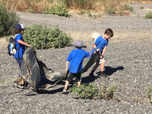 Three children hauling a big piece of trash out of a gravel creek bed