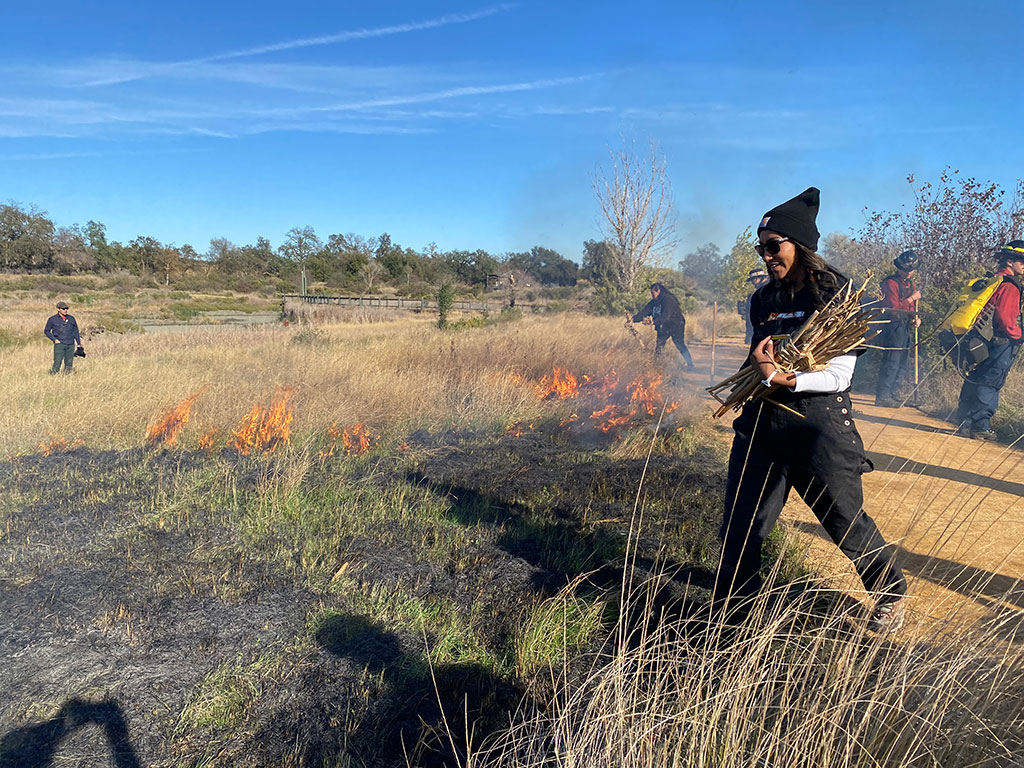 Melinda Adams carrying a tule bundle with a burning field in the background