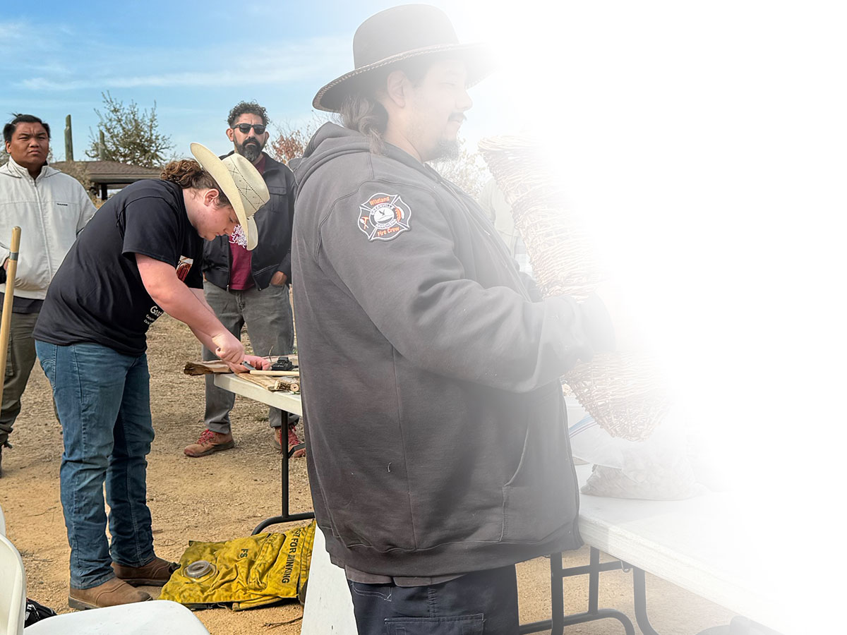 Young man in cowboy hat preparing to demonstrate how to start a fire by hand, while older men surround the demo table in support.