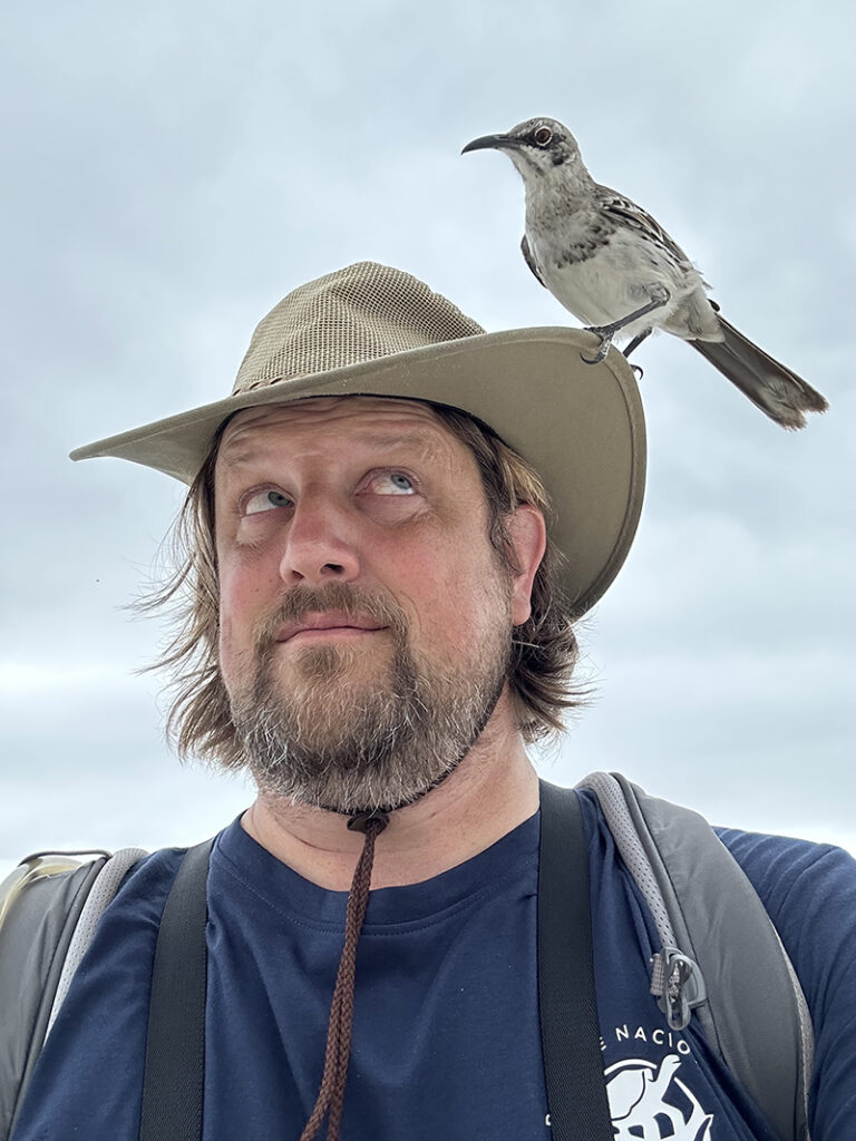 Bruce Christensen, a bearded man looking up at his safari hat where an Espanola Mockingbird is perched. 