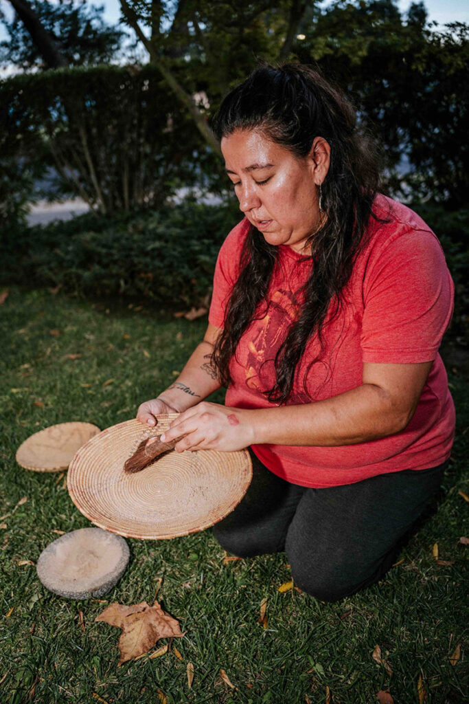 A CA Native woman, Christina Almendariz, kneeling on the grass working with basket weaving materials