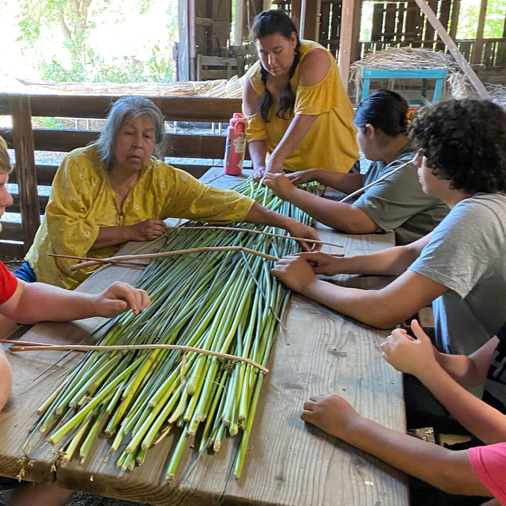 Diana and Christina Almendariz surrounded by students at a picnic table showing them how to weave tlaka mats